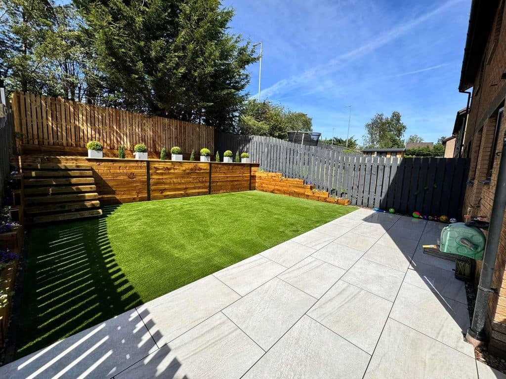 A garden with a grey porcelain patio, a neat lawn and a raised sleeper bed planted with clipped shrubs.