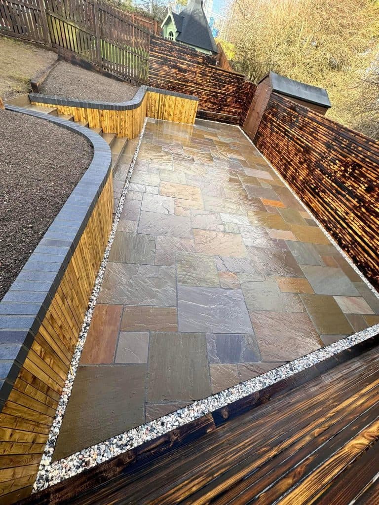 An Indian sandstone patio in mixed autumn tones, edged with pebbles and framed by timber sleepers and dark fencing.