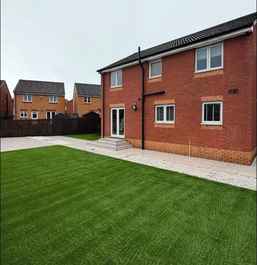 A wide striped lawn meeting a pale paved patio and stepped entrance at the back of a new-build house.