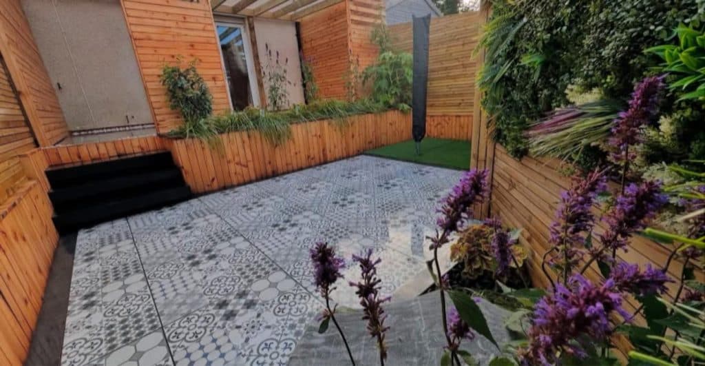 A small courtyard laid with patterned grey tiles, edged by timber-clad walls and planted borders.