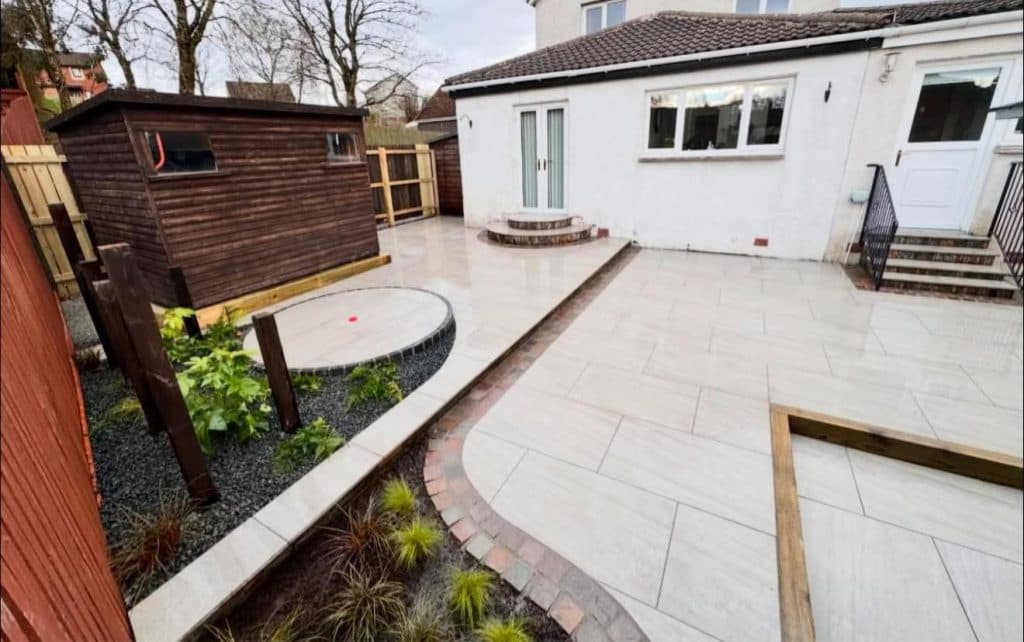 A pale porcelain patio and path curving around a planted border, with a circular paved base beside a timber shed.